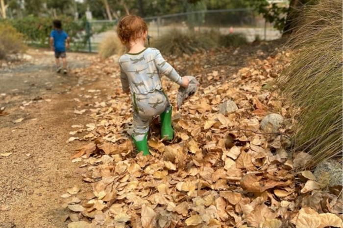 Small child in jammies and green boots walking through a pile of leaves