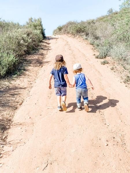 Two children, holding hands, walking up a trail surrounded by brush